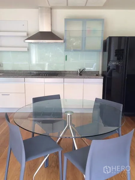 Condominium for rent in Khlong Toei, Bangkok - Modern open kitchen with white cabinets, range hood, black refrigerator, and a round glass dining table.