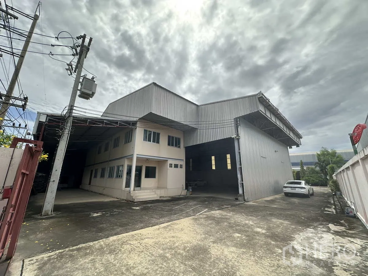 Factory for rent in Pathum Thani - Gated factory exterior with metal-clad warehouse, attached two-storey office and wide concrete driveway.