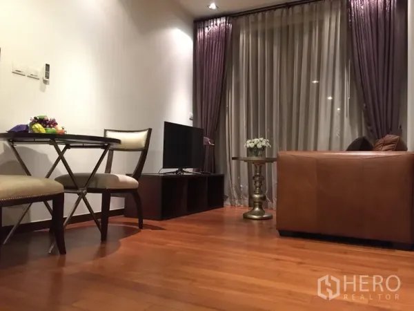 公寓 出租在Khlong Toei, Bangkok - 客厅配皮沙发、电视柜与小型餐桌，木地板与落地窗帘。