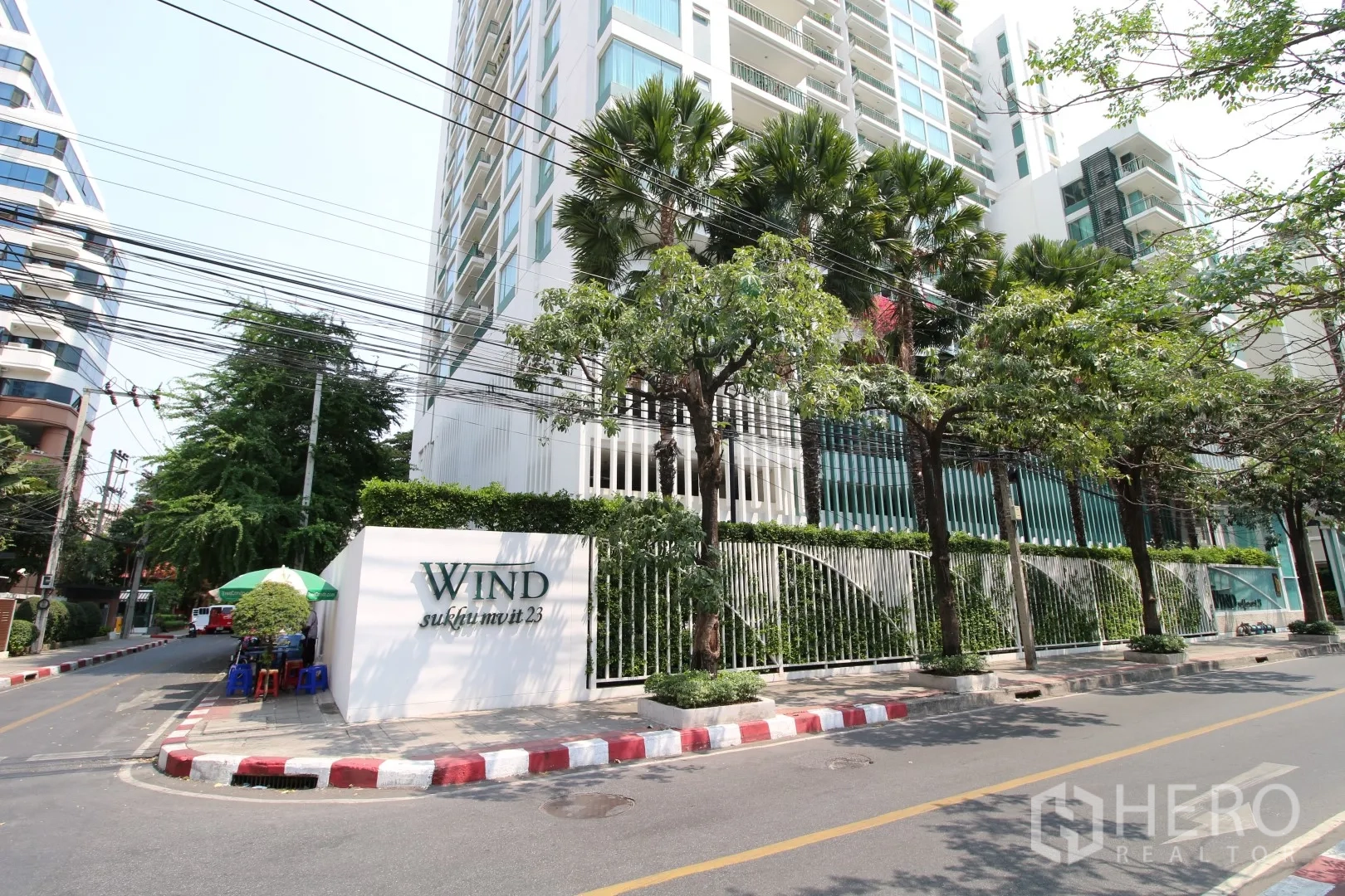 คอนโด ให้เช่าหรือขายในWatthana, Bangkok - ด้านหน้าอาคารคอนโดสมัยใหม่พร้อมรั้วและต้นปาล์มริมถนนเงียบสงบ