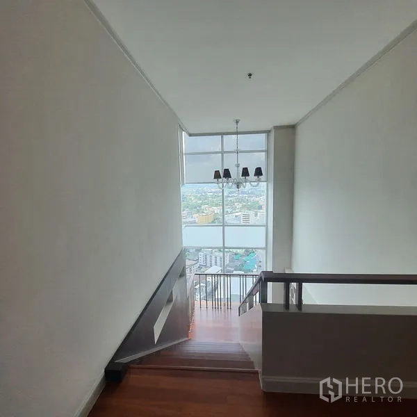 公寓 出租在Bang Kapi, Bangkok - 楼梯向下延伸至整面窗墙，配吊灯与城市景观。