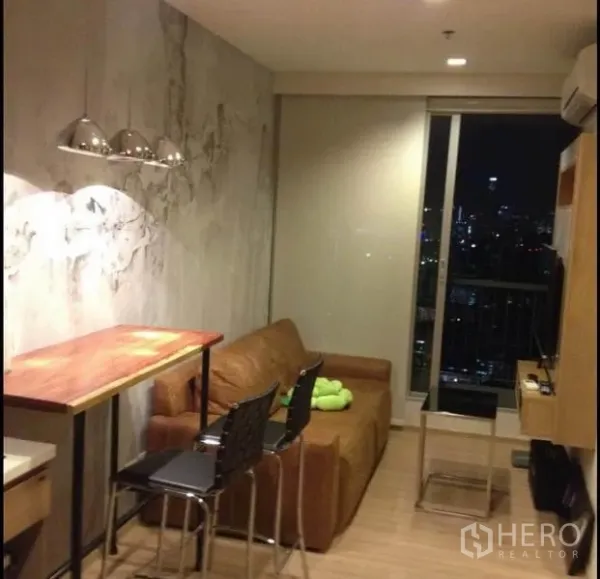 คอนโด ขายในPhaya Thai, Bangkok - ห้องนั่งเล่นโทนอบอุ่น โซฟาสีน้ำตาล โต๊ะบาร์และโคมไฟ หันสู่หน้าต่างเต็มบานวิวเมืองยามค่ำ