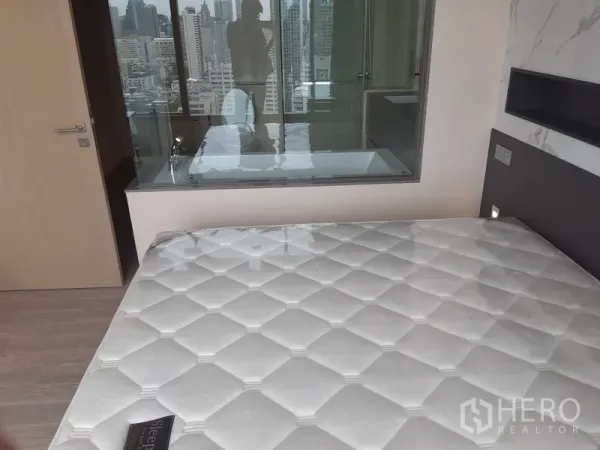 Condominium for rent or sale in Watthana, Bangkok - Bedroom looking toward glass-partition bathroom with city skyline beyond.