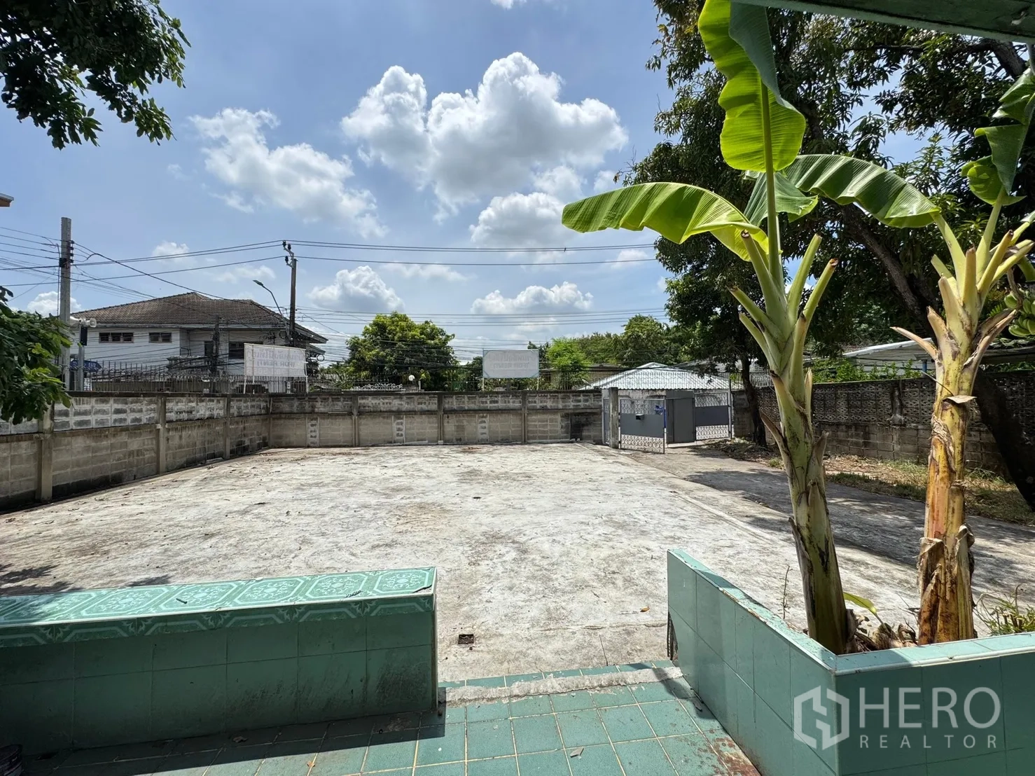 独栋房屋 for rent in Mueang Samut Prakan, Samut Prakan - 带围墙的大型混凝土庭院，绿色瓷砖台阶与香蕉树，在蓝天白云下。