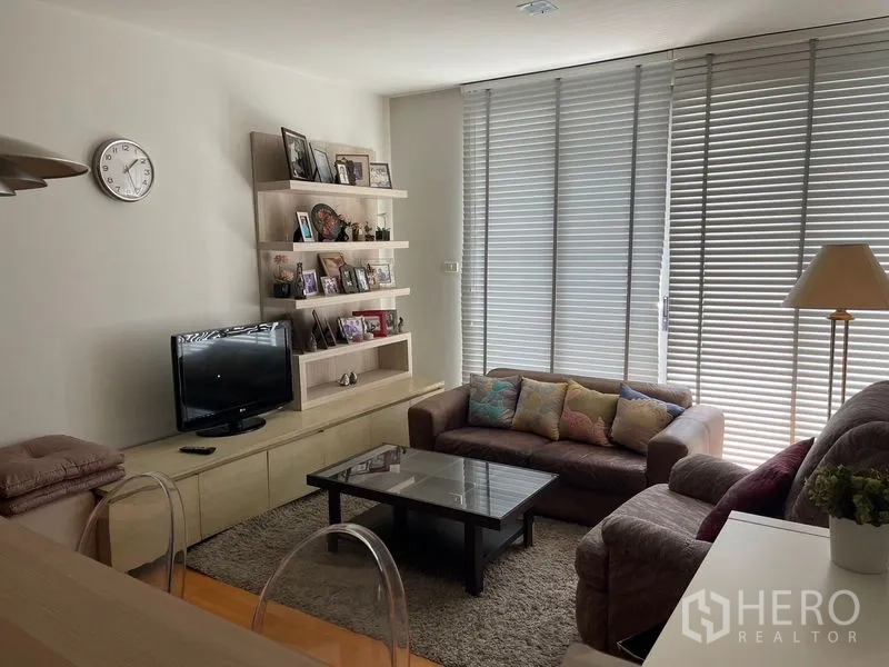 Condominium for sale in Khlong Toei, Bangkok - Living area with sofa set, TV console and display shelving facing tall windows.