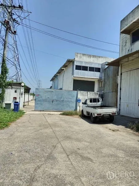 工厂 出售在Sai Noi, Nonthaburi - 工厂入口，金属大门、白色皮卡与宽阔混凝土车道，旁有高压线。