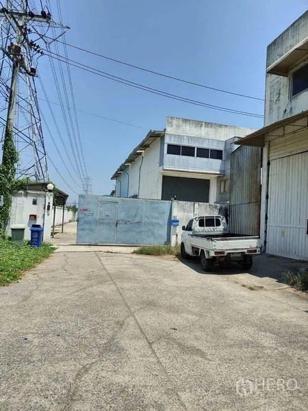 Factory for sale in Sai Noi, Nonthaburi - Gated factory entrance with a white pickup and wide concrete driveway under power lines.