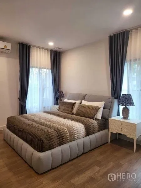 Detached House for rent in Saphan Sung, Bangkok - Cozy bedroom with upholstered bed, hardwood floor and tall curtained windows.