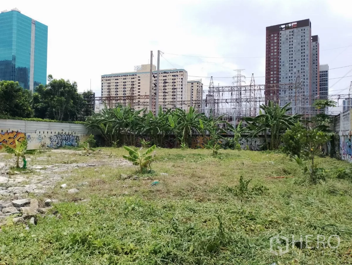Land for sale in Suan Luang, Bangkok - Cleared grassy site with boundary walls, graffiti art, and city buildings and power structures in the background.