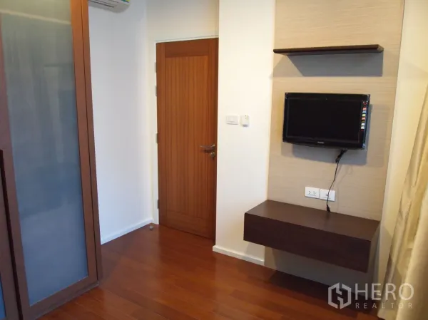 Condominium for rent or sale in Watthana, Bangkok - Second bedroom with wall-mounted TV, shelf and wooden door in warm tones.