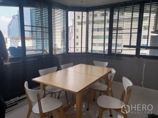 Condominium for rent in Khlong Toei, Bangkok - Bright dining area with a wooden table, six white chairs, and wraparound windows showing city views through blinds.