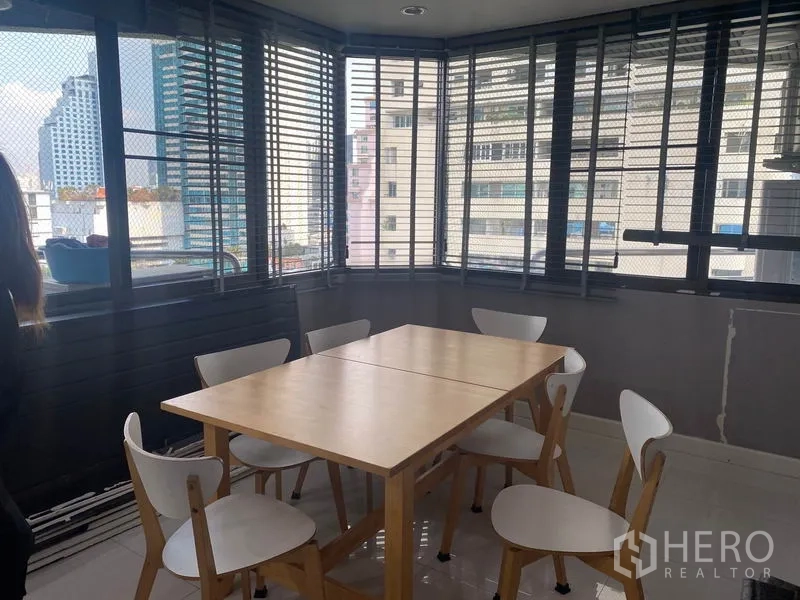Condominium for rent in Khlong Toei, Bangkok - Bright dining area with a wooden table, six white chairs, and wraparound windows showing city views through blinds.