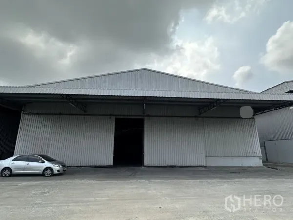คลังสินค้า ให้เช่าในMueang Samut Prakan, Samut Prakan - ด้านหน้าโกดังผนังเหล็กพร้อมประตูบานเลื่อนกว้าง ลานคอนกรีตและรถยนต์ ใต้ท้องฟ้าครึ้ม.