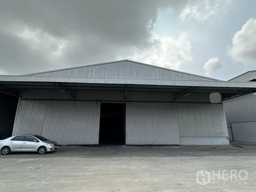 Warehouse for rent in Mueang Samut Prakan, Samut Prakan - Front view of a large steel‑clad warehouse with wide sliding doors and a car parked on the concrete forecourt under cloudy skies.