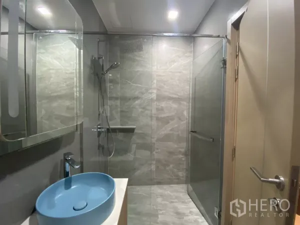 Condominium for rent or sale in Watthana, Bangkok - Modern bathroom with glass-enclosed rain shower, gray stone tiles and a blue vessel sink.