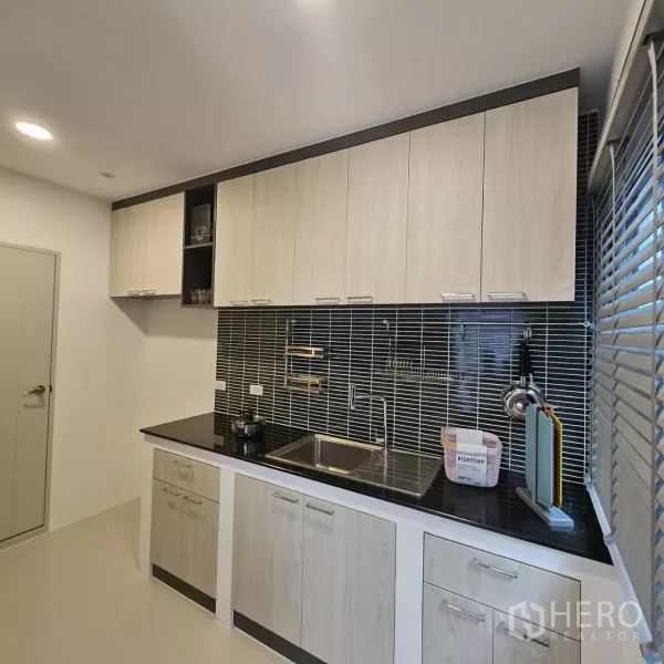 独立式住宅 出租在Bang Phli, Samut Prakan - 现代厨房，浅木色柜体，黑色台面与细条纹墙面瓷砖，旁有窗户。