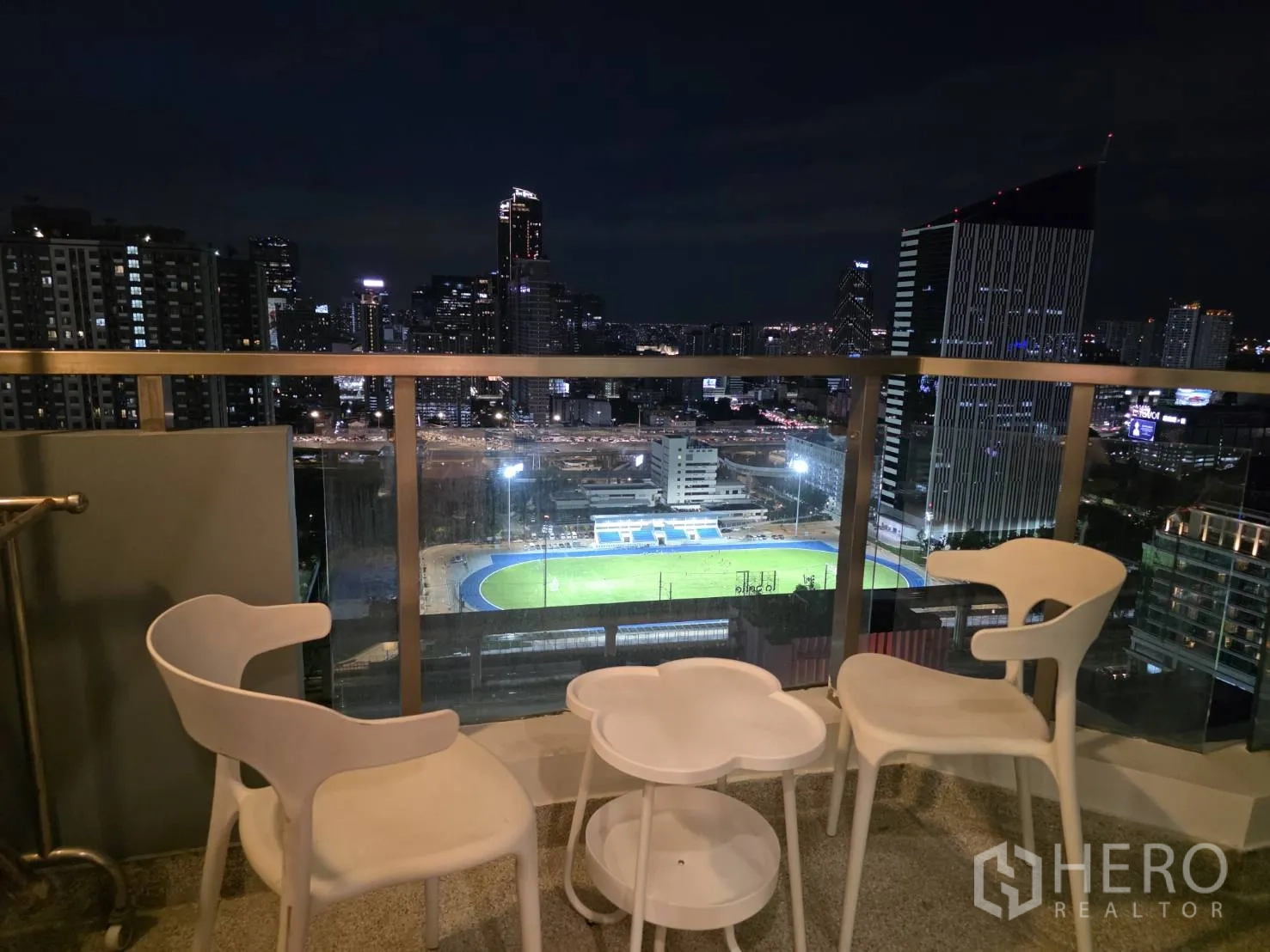 คอนโด for rent in Huai Khwang, Bangkok - ระเบียงยามค่ำพร้อมเก้าอี้สีขาวสามตัวและโต๊ะเล็ก มองเห็นแสงสีของเมือง
