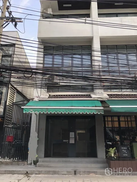 สำนักงาน ให้เช่าในWatthana, Bangkok - อาคารพาณิชย์สูงสามชั้น ด้านหน้าเป็นผ้าใบกันสาดสีเขียว ประตูกระจก ช่องหน้าต่างเหล็กดัด และสายไฟเหนือถนนในเมือง