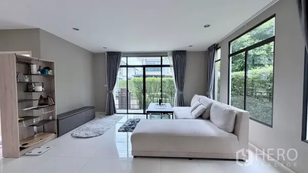 บ้าน ให้เช่าหรือขายในBang Phli, Samut Prakan - ห้องนั่งเล่นสว่างผนังกระจก ผ้าม่านสีเทา และโซฟา L‑shape พร้อมโต๊ะกลาง