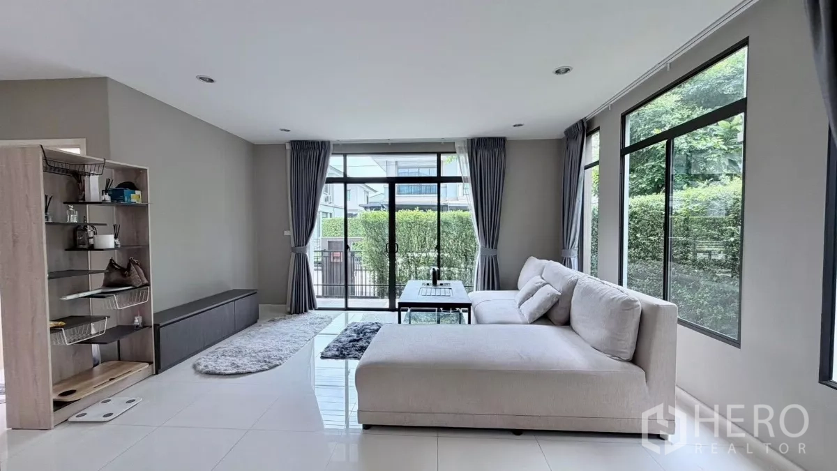 บ้าน ให้เช่าหรือขายในBang Phli, Samut Prakan - ห้องนั่งเล่นสว่างผนังกระจก ผ้าม่านสีเทา และโซฟา L‑shape พร้อมโต๊ะกลาง