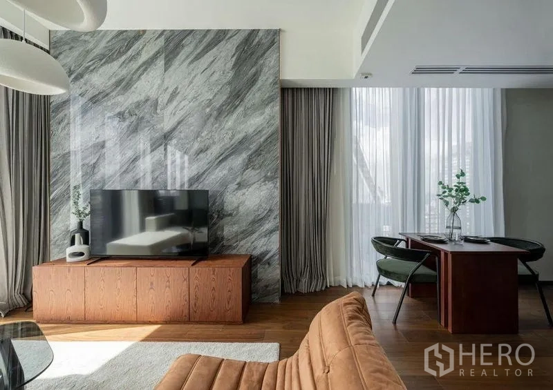 Condominium for rent in Khlong Toei, Bangkok - TV console against a marble accent wall with a dining corner and sheer curtains in the living area.