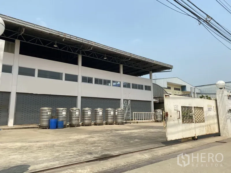 Factory for sale in Khlong Luang, Pathum Thani - Wide gated entrance and loading yard facing multiple roller shutter bays.