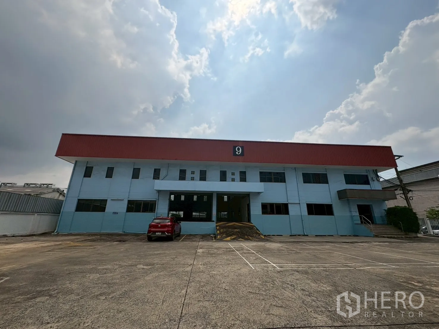 คลังสินค้า for rent in Phra Khanong, Bangkok - ด้านหน้าโกดังสองชั้นสีฟ้าพร้อมหลังคาสีแดงและลานคอนกรีตกว้าง ใต้ท้องฟ้ามีเมฆ.