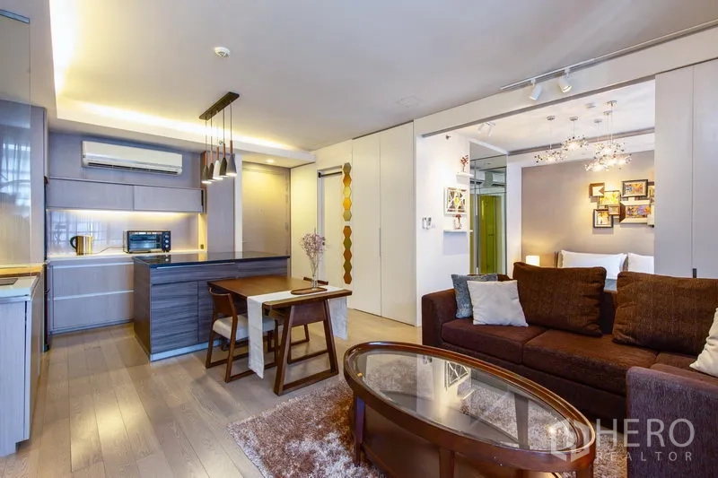 Condominium for rent in Watthana, Bangkok - Open-plan living room with brown sofa, glass coffee table, dining set, and modern kitchen island with pendant lights.