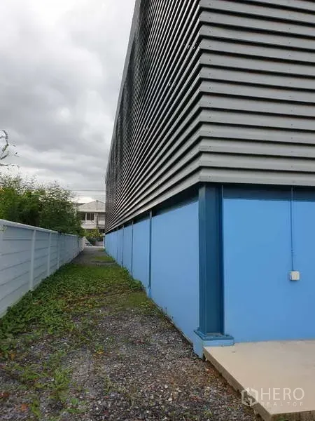 Factory for sale in Sai Noi, Nonthaburi - Side view showing corrugated metal cladding over a blue base wall along the fenced boundary of the factory.