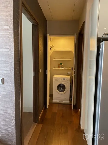 Condominium for rent in Watthana, Bangkok - Compact laundry closet off the corridor with front‑loading washing machine and storage shelves.