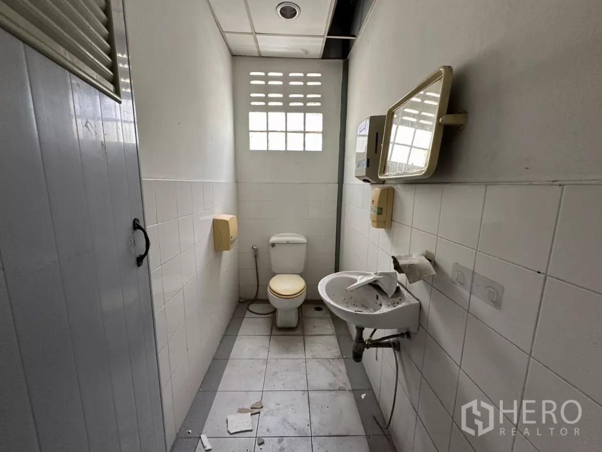 Warehouse for rent in Suan Luang, Bangkok - Second restroom with window, white tiles, toilet, and compact wall-mounted sink.