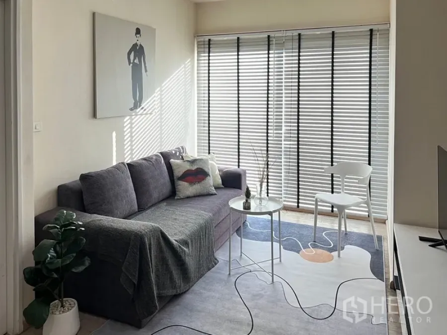 Condominium for rent in Khlong Toei, Bangkok - Sunlit living area with grey sofa, wall art, blinds over full-height windows, and a small coffee table.