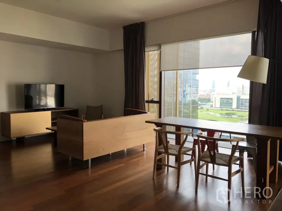 Condominium for rent in Pathum Wan, Bangkok - Open-plan living and dining room with hardwood floors, TV console, and floor-to-ceiling windows overlooking city and green park views.