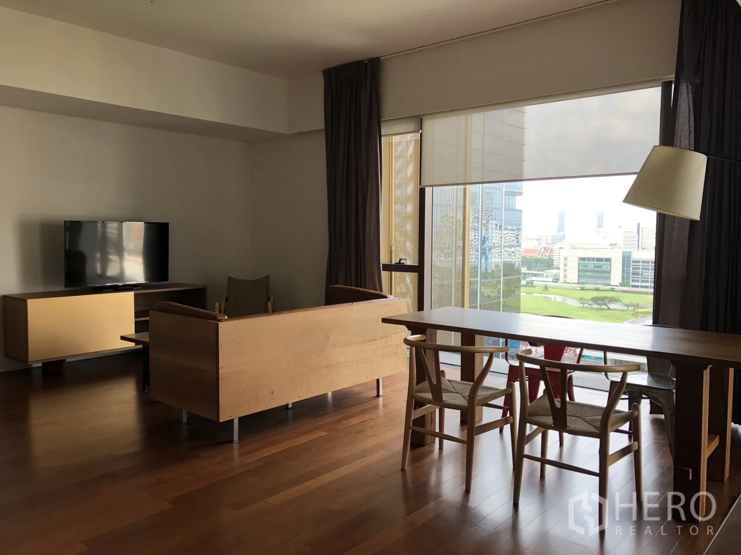 Condominium for rent in Pathum Wan, Bangkok - Open-plan living and dining room with hardwood floors, TV console, and floor-to-ceiling windows overlooking city and green park views.