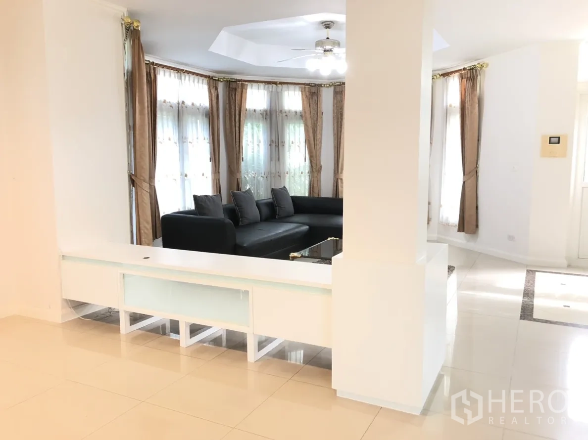Detached House for rent or sale in Lat Phrao, Bangkok - Open-plan lounge with tiled floors, a white built-in console and large curtained windows.