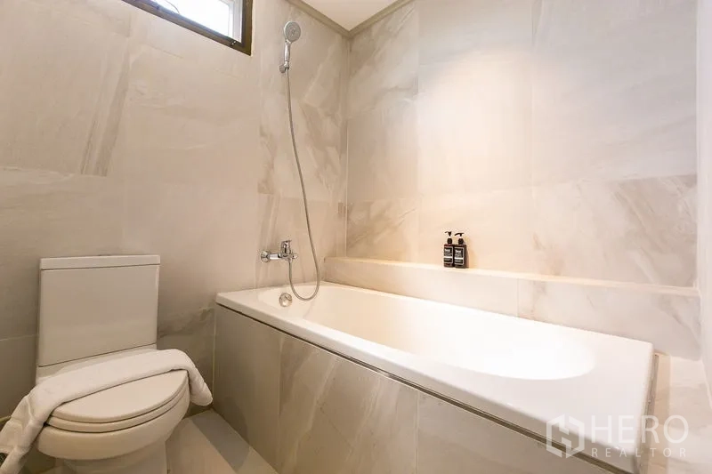 Apartment for rent in Watthana, Bangkok - Modern bathroom with bathtub and shower, light marble-style tiles and toilet.