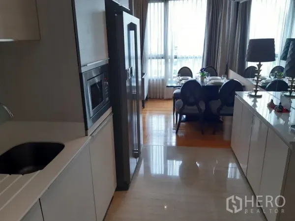 公寓 出租在Watthana, Bangkok - 走廊式厨房，白色橱柜、嵌入式微波炉与双门冰箱，通向采光充足的餐区和落地窗。