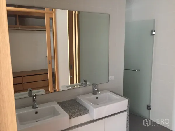 Condominium for rent in Khlong Toei, Bangkok - Contemporary bathroom featuring double vanity sinks, large mirror and a frosted-glass shower next to a walk-in wardrobe.