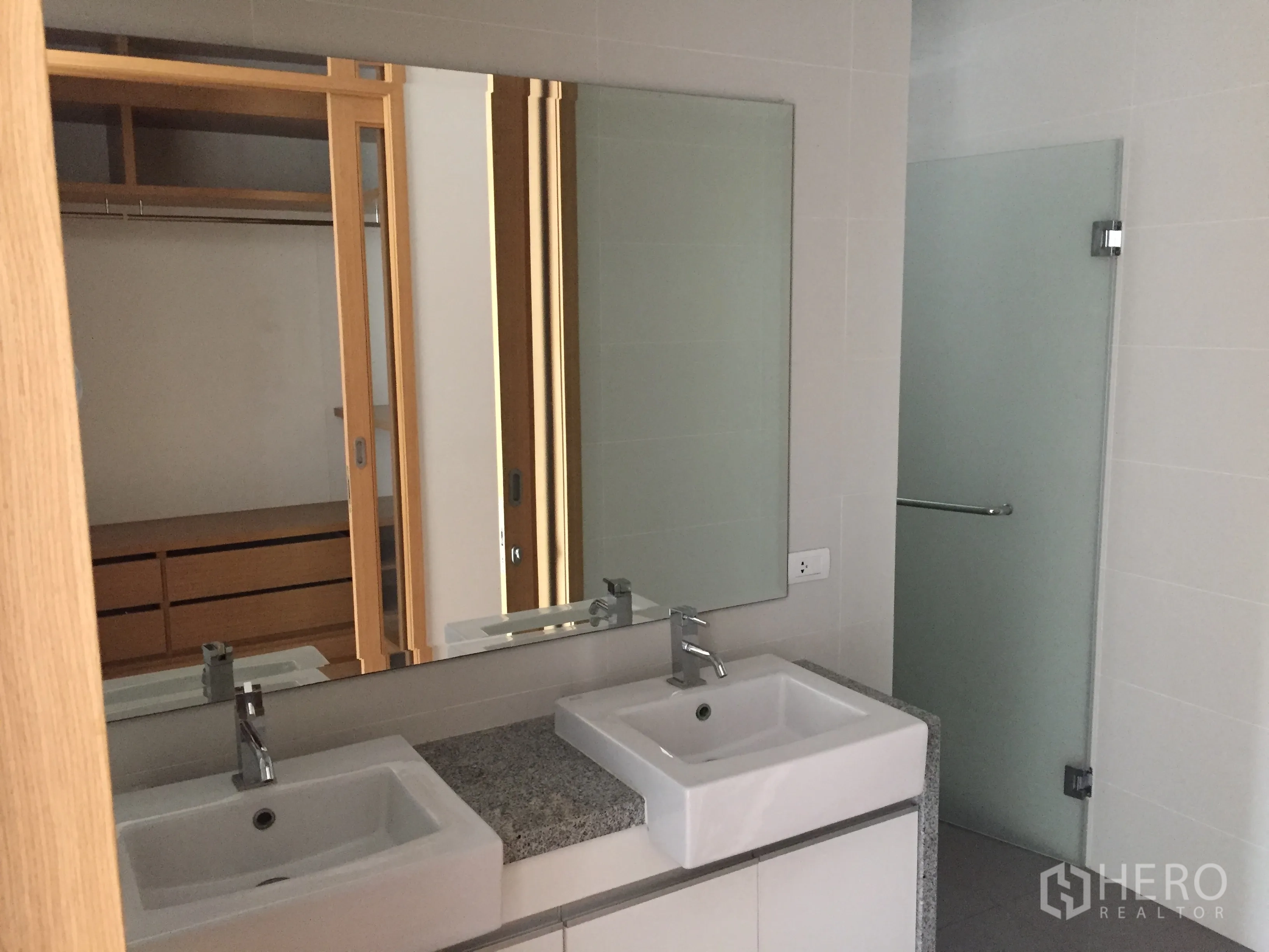 Condominium for rent in Khlong Toei, Bangkok - Contemporary bathroom featuring double vanity sinks, large mirror and a frosted-glass shower next to a walk-in wardrobe.