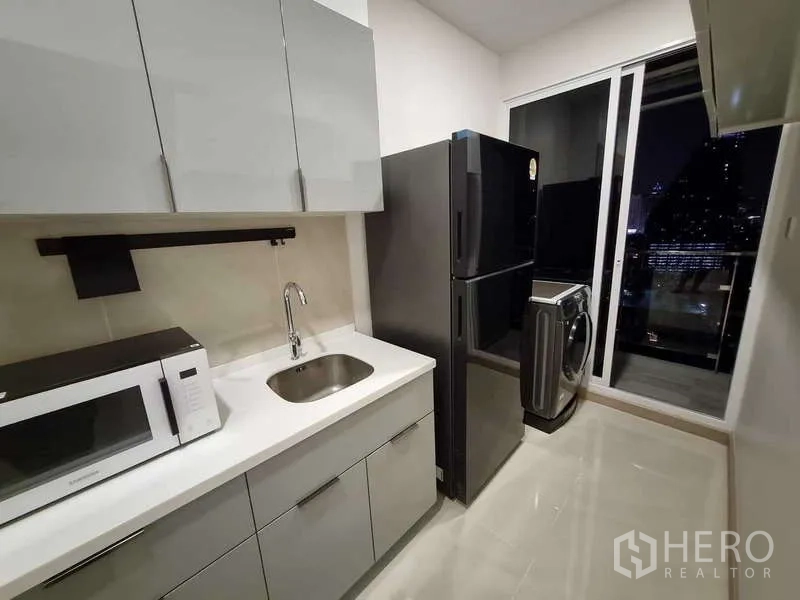 Condominium for rent - Modern galley kitchen with grey cabinets, fridge and washer beside a balcony with night city view.