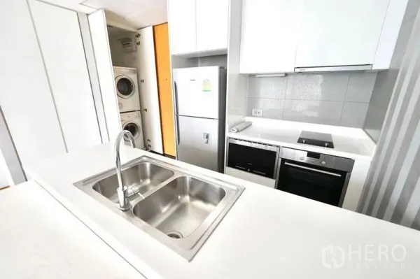 Condominium for rent or sale in Watthana, Bangkok - Compact kitchen featuring double sink, cooktop, oven, fridge and a stacked washer-dryer in a cabinet.