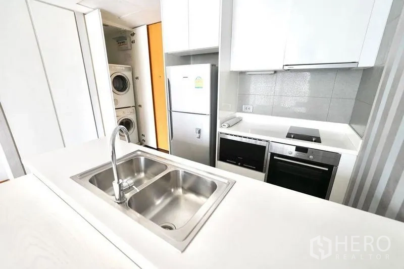 Condominium for rent or sale in Watthana, Bangkok - Compact kitchen featuring double sink, cooktop, oven, fridge and a stacked washer-dryer in a cabinet.