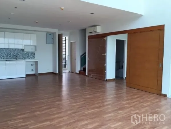 Condominium for sale in Bang Na, Bangkok - Open-plan living and dining space with a compact white kitchen, wood flooring and sliding wooden doors.