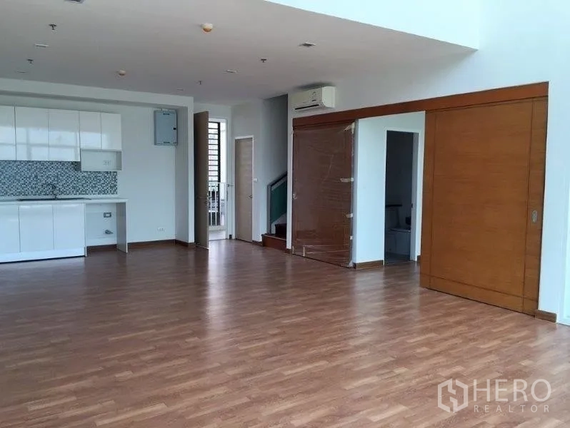 Condominium for sale in Bang Na, Bangkok - Open-plan living and dining space with a compact white kitchen, wood flooring and sliding wooden doors.