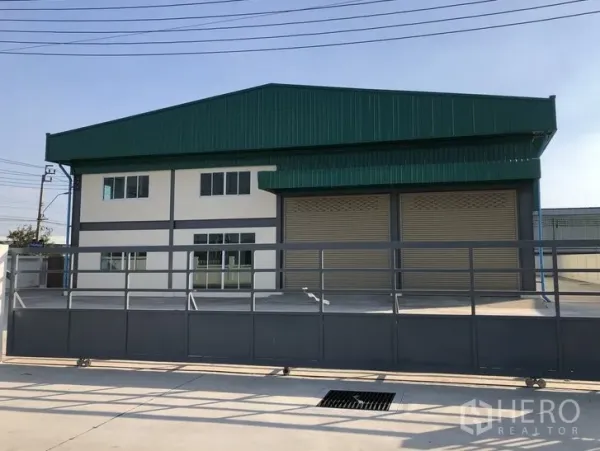 工厂 出租在Mueang Samut Prakan, Samut Prakan - 绿色金属屋顶的工厂外立面，设两扇卷帘门、两层办公室、宽阔混凝土前院与滑动大门