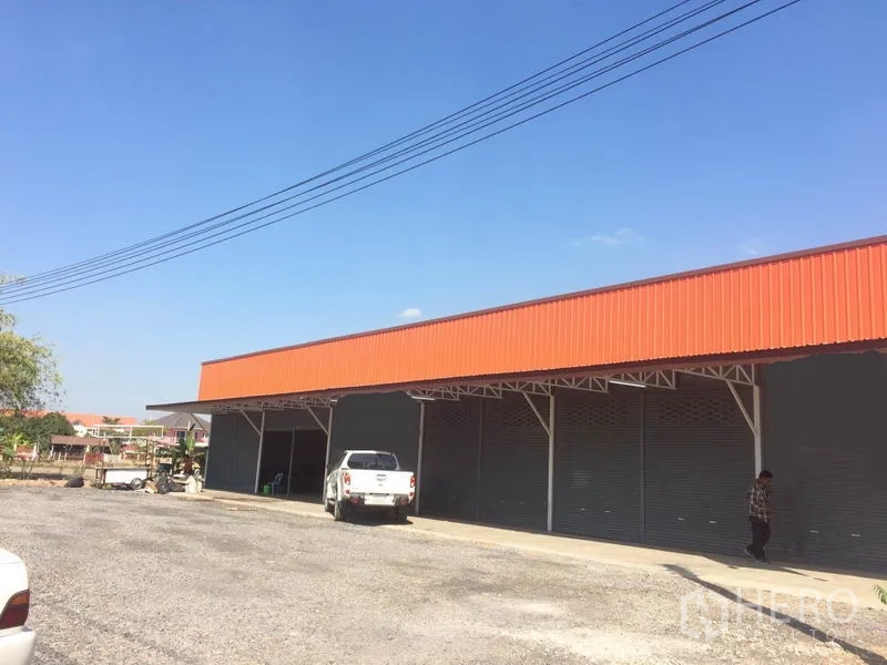 货栈 出租在Pathum Thani - 仓库外观，橙色山墙，多个卷帘门，前方有宽阔装卸与停车场地，位于兰鲁甲。