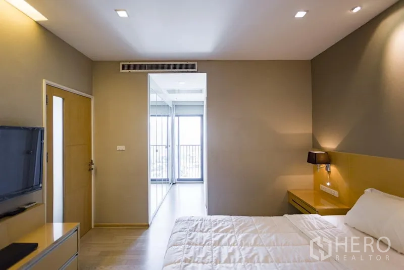 Condominium for rent in Khlong Toei, Bangkok - Bedroom view toward balcony with mirrored wardrobe, bedside lighting and floor-to-ceiling window.