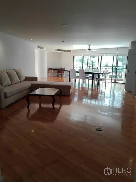 Condominium for rent in Khlong Toei, Bangkok - Expansive open-plan living room with polished wood flooring, sofa, coffee table, and full-height windows opening to a balcony.