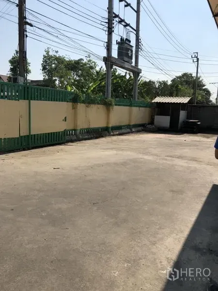 Warehouse for rent in Bueng Kum, Bangkok - Gated concrete yard beside the warehouse with perimeter wall, power lines, and a small guard hut.