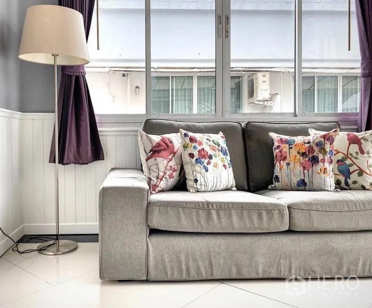 Condominium for rent in Khlong Toei, Bangkok - Close-up of gray fabric sofa with floral pillows beside window and floor lamp.
